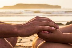 Zwei Menschen halten einander sanft die Hände am Strand bei Sonnenuntergang, im Hintergrund das Meer und eine Insel.